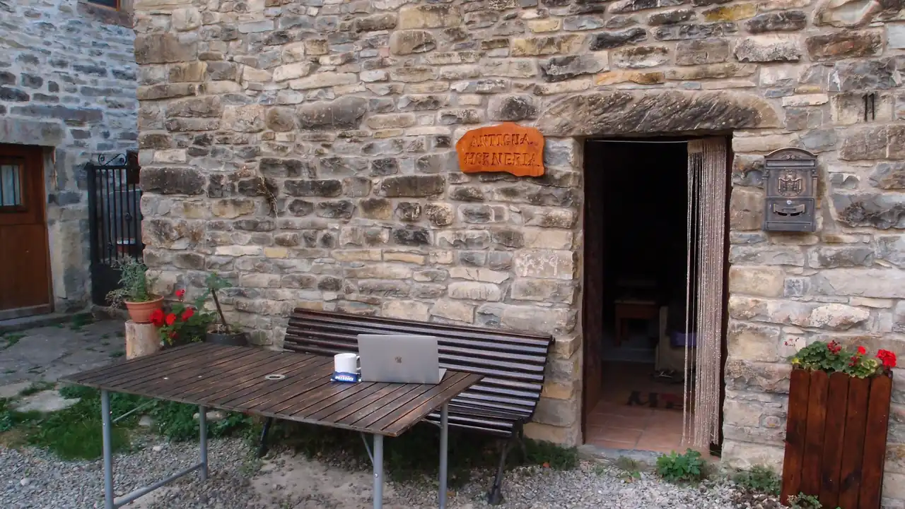 A stone courtyard in the Pyrenees: an old bakery building, an outdoor table with a laptop and a mug